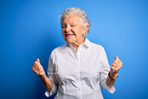 Старша красива жінка в елегантній сорочці, що стоїть над ізольованим синім тлом, збуджена успіхом з піднятими руками і закритими очима, що святкують перемогу посміхаючись. концепція переможця
. - Фото, зображення