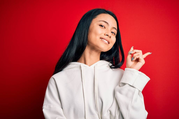 Young beautiful chinese sporty woman wearing sweatshirt over isolated red background with a big smile on face, pointing with hand and finger to the side looking at the camera. - 写真・画像