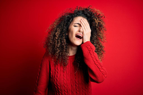 Young beautiful woman with curly hair and piercing wearing casual red sweater Yawning tired covering half face, eye and mouth with hand. Face hurts in pain. - Photo, Image
