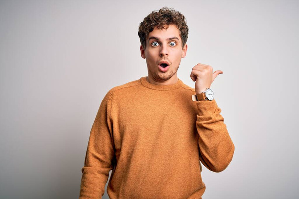Young blond handsome man with curly hair wearing casual sweater over white background Surprised pointing with hand finger to the side, open mouth amazed expression. - Foto, immagini