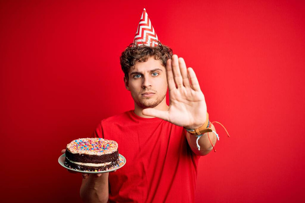 Mladý blonďatý muž s kudrnatými vlasy na hlavě narozeninový klobouk drží dort nad červeným pozadím s otevřenou rukou dělá stop znamení s vážným a sebejistý výraz, obrana gesto - Fotografie, Obrázek