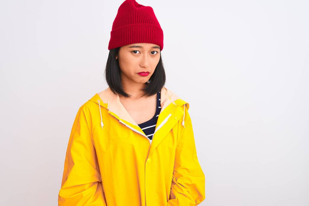 Νεαρή όμορφη Κινέζα γυναίκα φορώντας αδιάβροχο και μάλλινο καπέλο πάνω από απομονωμένο λευκό φόντο κατάθλιψη και να ανησυχείτε για την αγωνία, κλάμα θυμωμένος και φοβισμένος. Θλιβερή έκφραση. - Φωτογραφία, εικόνα