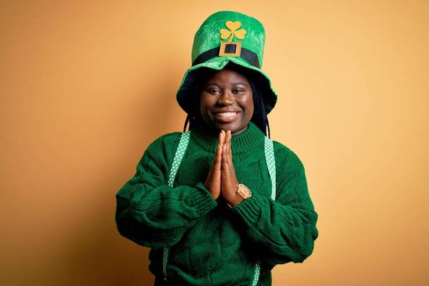 Συν μέγεθος αφροαμερικανή γυναίκα με κοτσίδες φορώντας πράσινο καπέλο με τριφύλλι την ημέρα του Αγίου Πατρικίου προσεύχεται με τα χέρια μαζί ζητώντας συγχώρεση χαμογελώντας σίγουροι. - Φωτογραφία, εικόνα