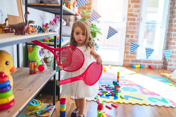 Adorable blonde toddler playing tennis using racket and ball around lots of toys at kindergarten - Foto, Imagen