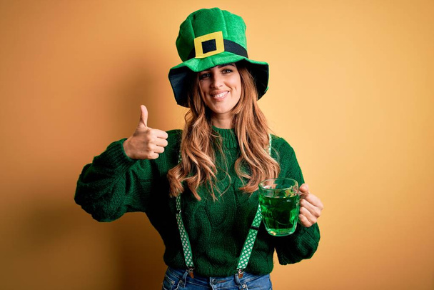 Beautiful woman wearing hat drinking jar of green beverage celebrating saint patricks day happy with big smile doing ok sign, thumb up with fingers, excellent sign - Foto, immagini