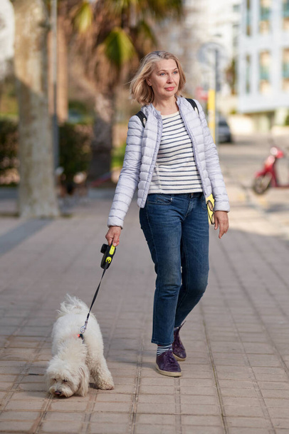Mulher loira de meia-idade atraente andando com cachorro branco fofo na cidade de verão
 - Foto, Imagem