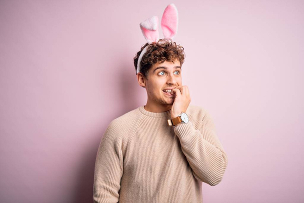 Jeune homme blond beau portant des oreilles de lapin de Pâques sur fond rose isolé regardant stressé et nerveux avec les mains sur les ongles mordants de la bouche. Problème d'anxiété
. - Photo, image
