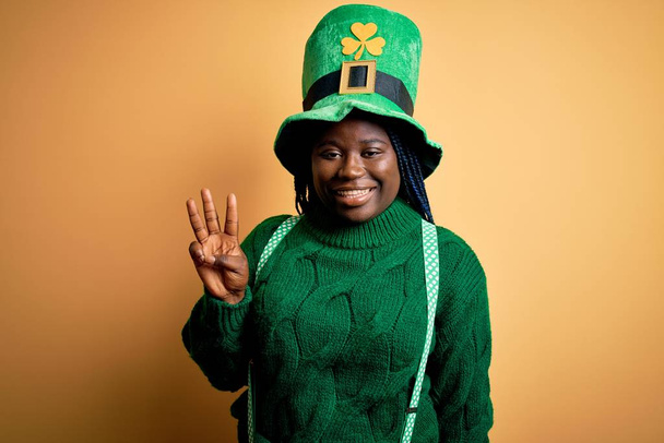 Плюс размер африканский американец женщина с косичками носить зеленую шляпу с клевером в день Святого Патрика показывая и указывая пальцами номер три улыбаясь уверенно и счастливо
. - Фото, изображение