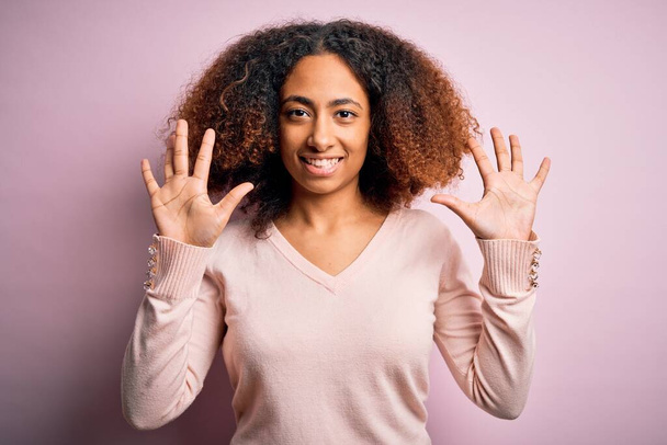 Afro saçlı, pembe arka planda günlük süveter giyen ve parmaklarını on numarayı gösteren, kendinden emin ve mutlu gülümseyen Afro Amerikalı genç bir kadın.. - Fotoğraf, Görsel