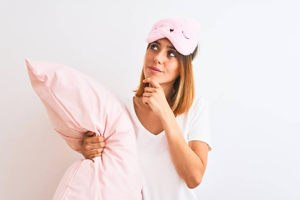 Krásná zrzka žena nosí spací masku mazlení růžový polštář přes izolované pozadí vážná tvář přemýšlení o otázce, velmi zmatený nápad - Fotografie, Obrázek