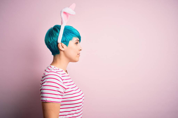 Jeune femme aux cheveux bleus mode portant des oreilles de lapin de Pâques sur fond rose regardant vers le côté, pose de profil relax avec visage naturel avec sourire confiant
. - Photo, image