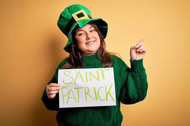 Mujer de talla grande con sombrero verde celebrando el día de San Patricio sosteniendo el estandarte del día de San Patricio muy feliz señalando con la mano y el dedo hacia un lado
 - Foto, Imagen