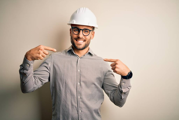 Jonge architect man draagt bouwer veiligheidshelm over geïsoleerde achtergrond kijken zelfverzekerd met glimlach op het gezicht, wijzend zichzelf met vingers trots en gelukkig. - Foto, afbeelding