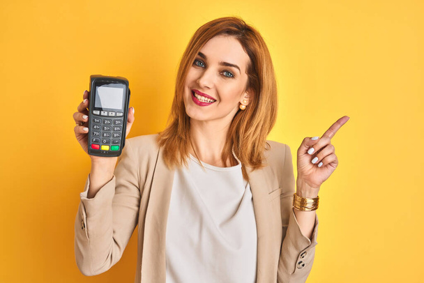 Redhead caucasian business woman holding point of sale terminal over isolated background very happy pointing with hand and finger to the side - Фото, зображення
