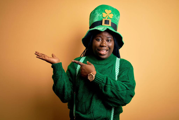 Африканская женщина plus size с брейдами в зеленой шляпе с клевером в день святого Патрика удивляла и улыбалась фотоаппарату, показывая рукой и показывая пальцем
. - Фото, изображение