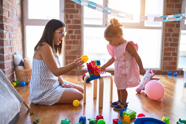 Young beautiful teacher and toddler playing with building blocks toy on the table at kindergarten - Photo, Image