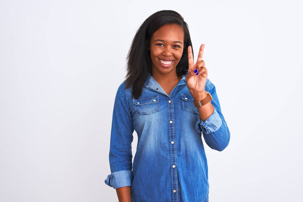 Mooie jonge Afro-Amerikaanse vrouw draagt spijkerjasje over geïsoleerde achtergrond glimlachend kijkend naar de camera die vingers laat zien die een overwinningsteken doen. Nummer twee. - Foto, afbeelding