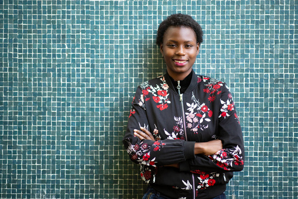 Retrato frontal de una joven africana sonriente con los brazos cruzados por el fondo de mosaico
 - Foto, Imagen