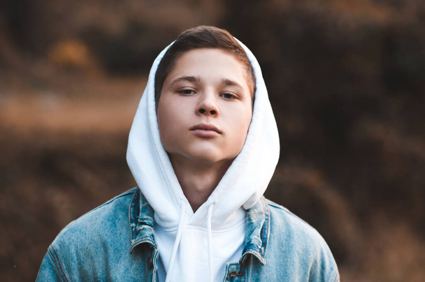 Tiener jongen 16-17 jaar oud draagt witte hoodie en spijkerjasje close-up. Ik kijk naar de camera. Twintiger jaren.  - Foto, afbeelding
