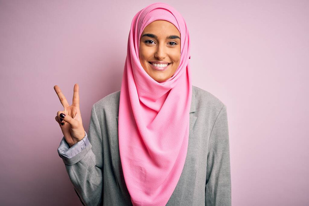 Joven hermosa mujer de negocios morena vistiendo hijab musulmán rosa y chaqueta de negocios sonriendo con la cara feliz guiñando el ojo a la cámara haciendo signo de victoria. Número dos.
. - Foto, imagen