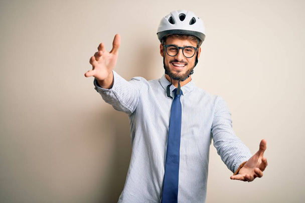 孤立した白い背景の上に立っている眼鏡や自転車のヘルメットを身に着けている若いビジネスマンは、抱擁のためのオープンアームで笑顔カメラを見て。幸せを受け入れる陽気な表情. - 写真・画像