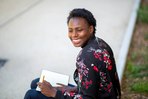 Close up lato ritratto felice giovane donna africana seduta fuori a scrivere nel libro
  - Foto, immagini