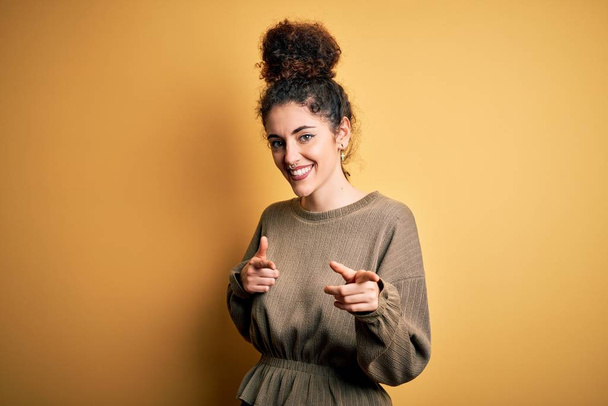 Young beautiful brunette woman with curly hair and piercing wearing casual dress pointing fingers to camera with happy and funny face. Good energy and vibes. - Photo, Image