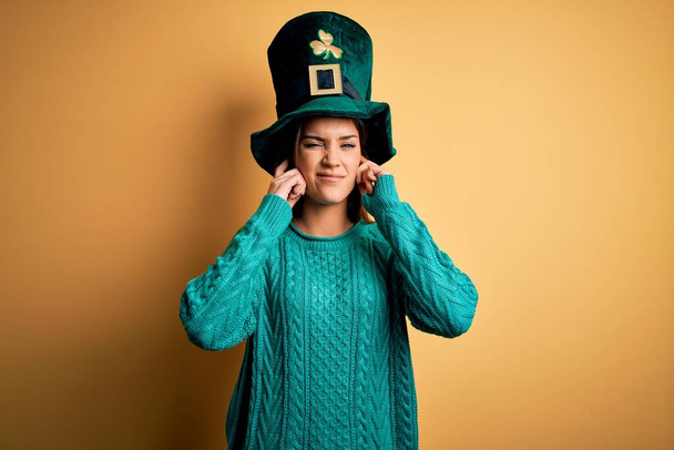 Jeune belle femme brune portant un chapeau vert avec trèfle célébrant saint patricks jour couvrant les oreilles avec les doigts avec une expression agacée pour le bruit de la musique forte. Concept sourd
. - Photo, image