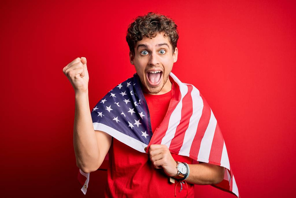 Mladý blonďatý patriotický muž s kudrnatými vlasy na sobě má vlajku USA, oslavující den nezávislosti, křičící hrdě a oslavující vítězství a úspěch, velmi vzrušený, jásající emoce - Fotografie, Obrázek