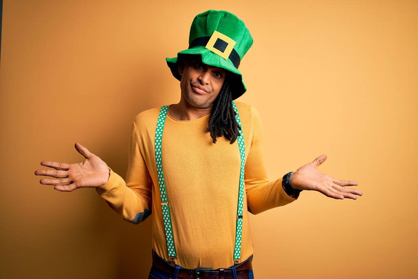 Joven afroamericano vestido con sombrero verde celebrando el día de santo patricks expresión despistada y confusa con los brazos y las manos levantadas. Concepto de duda
. - Foto, imagen