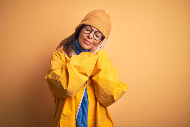 Middle age woman wearing yellow raincoat and winter hat over isolated background sleeping tired dreaming and posing with hands together while smiling with closed eyes. - 写真・画像