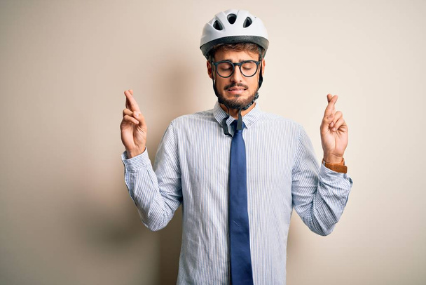 孤立した白いベークグラウンドのジェスチャー指の上に立つ眼鏡や自転車のヘルメットを身に着けている若いビジネスマンは、希望と目を閉じて笑顔を越えました。幸運と迷信の概念. - 写真・画像