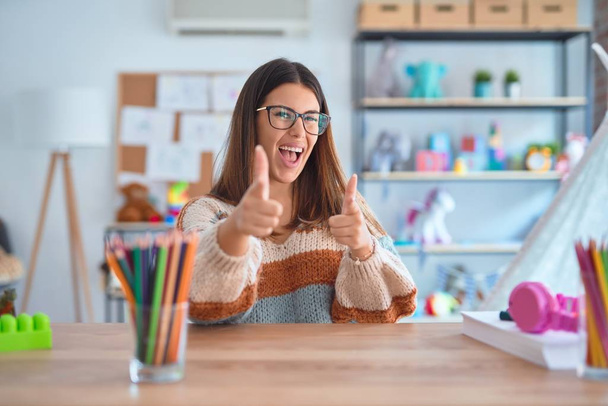 Młoda piękna nauczycielka w swetrze i okularach siedząca na biurku w przedszkolu wskazująca palcem na kamerę ze szczęśliwą i zabawną twarzą. Dobra energia i wibracje. - Zdjęcie, obraz