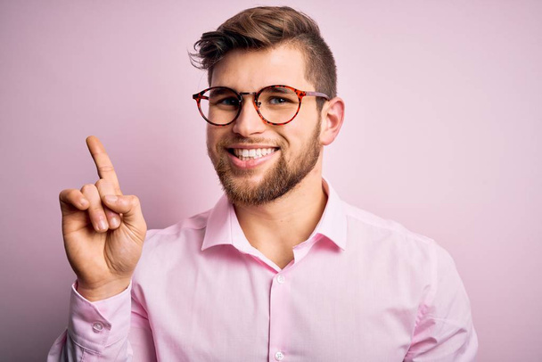 Mladý pohledný blonďatý muž s vousy a modrýma očima v růžové košili a brýlích ukazující a ukazující prstem číslo jedna a usmívající se sebevědomě a šťastně. - Fotografie, Obrázek