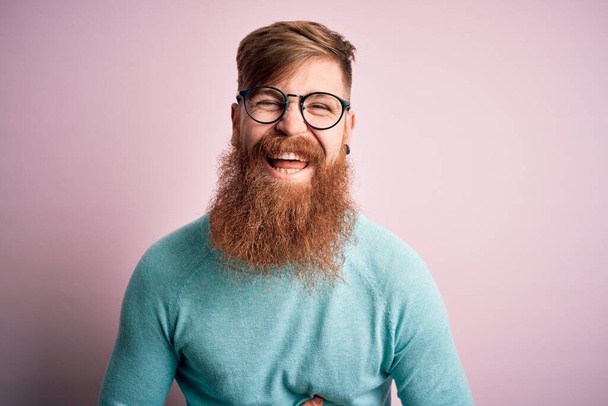 Jóképű ír vöröshajú férfi szakállal szemüveget visel rózsaszín elszigetelt háttér mosolyogva és nevetve hangosan, mert vicces őrült vicc kezekkel a testen. - Fotó, kép