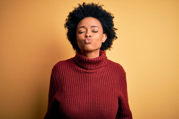 Young beautiful African American afro woman with curly hair wearing casual turtleneck sweater looking at the camera blowing a kiss on air being lovely and sexy. Love expression. - Photo, Image
