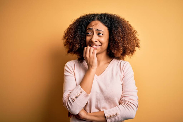 Young african american woman with afro hair wearing casual sweater over yellow background looking stressed and nervous with hands on mouth biting nails. Anxiety problem. - Photo, image