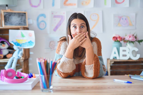 Young beautiful teacher woman wearing sweater and glasses sitting on desk at kindergarten shocked covering mouth with hands for mistake. Secret concept. - Photo, Image