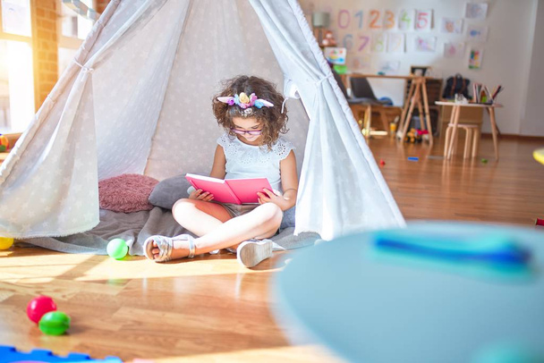 美しい幼児身に着けている眼鏡とユニコーンdiadem座って床の上にtipi読書本で幼稚園 - 写真・画像