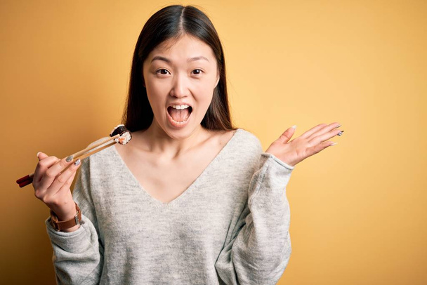Young asian woman eating japanese food, holding salmon and rice maki sushi using chopsticks very happy and excited, winner expression celebrating victory screaming with big smile and raised hands - Photo, image