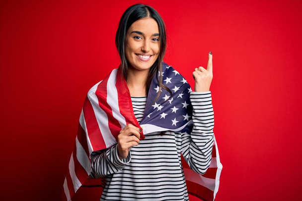 Jeune belle femme patriotique brune portant le drapeau des États-Unis pour la journée de l'indépendance surprise par une idée ou une question pointant du doigt avec un visage heureux, numéro un
 - Photo, image