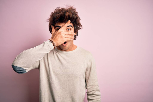 Jonge knappe man draagt casual t-shirt staan over geïsoleerde roze achtergrond gluren in shock bedekking gezicht en ogen met de hand, kijken door vingers met beschaamde uitdrukking. - Foto, afbeelding