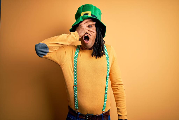 Mladý africký Američan v zeleném klobouku slaví svaté patricks den nakukuje v šoku zakrývající tvář a oči rukou, dívá se prsty s rozpačitým výrazem. - Fotografie, Obrázek