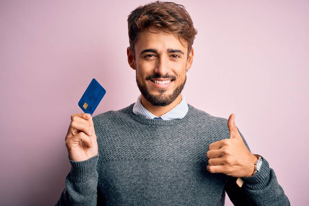 Молодой клиент с бородой держа кредитную карту для оплаты за розовый фон доволен большой улыбкой делает хорошо знак, палец вверх пальцами, отличный знак
 - Фото, изображение