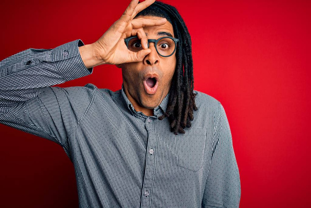 Mladý pohledný africký Američan s dredy v neformální košili a brýlích dělá v pořádku gesto šokované překvapeným obličejem, oči hledící skrz prsty. Nevěřící výraz. - Fotografie, Obrázek