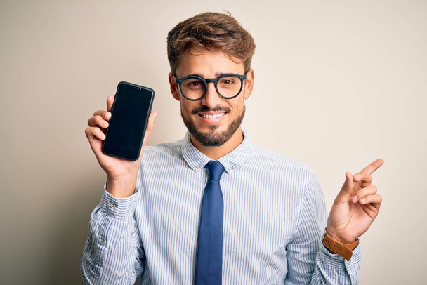 Молодой бизнесмен, держащий смартфон на изолированном белом фоне, очень доволен, указывая рукой и пальцем в сторону.
 - Фото, изображение