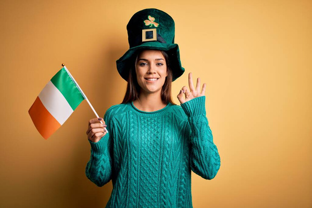 schöne Frau mit grünem Hut feiert den Heiligen Patricks Day mit irischer irischer Flagge tut ok Zeichen mit den Fingern, ausgezeichnetes Symbol - Foto, Bild