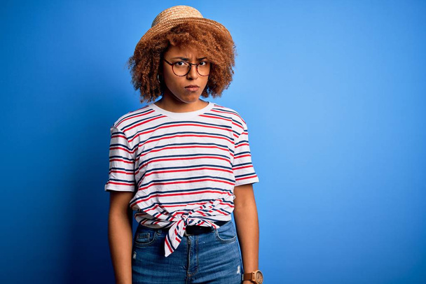 Αφροαμερικανή γυναίκα με σγουρά μαλλιά σε διακοπές φορώντας καλοκαιρινό καπέλο και ριγέ t-shirt σκεπτική και νευρική, συνοφρυωμένη λόγω προβλήματος. Αρνητικό πρόσωπο. - Φωτογραφία, εικόνα