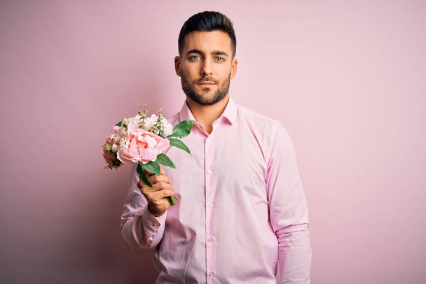 Νεαρός ρομαντικός άντρας που κρατάει μπουκέτο από ανοιξιάτικα λουλούδια πάνω από ροζ απομονωμένο φόντο με μια σίγουρη έκφραση στο έξυπνο πρόσωπο που σκέφτεται σοβαρά - Φωτογραφία, εικόνα