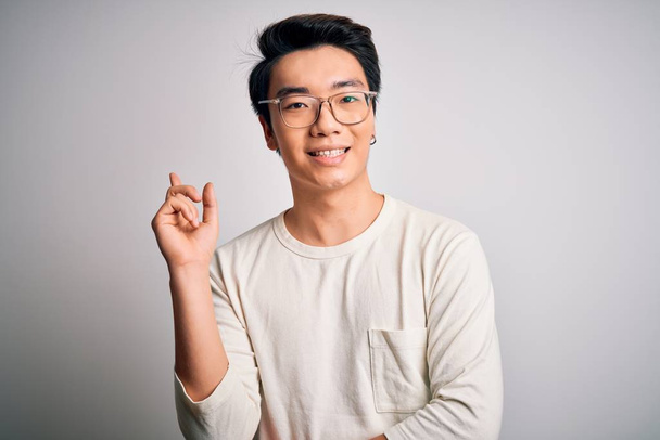 Jeune bel homme chinois portant un t-shirt décontracté et des lunettes sur fond blanc avec un grand sourire sur le visage, pointant avec la main et le doigt vers le côté en regardant la caméra
. - Photo, image